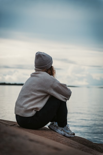 A person in a beanie and sweater sitting on a rock by the water