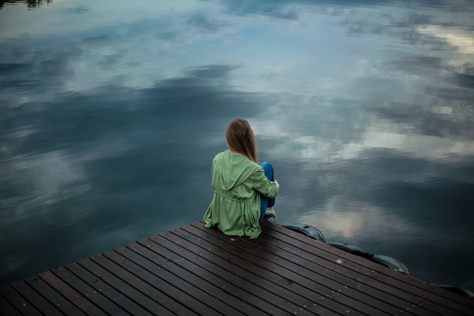 A woman with depression sits on a wooden dock, reflecting by a calm lake under a cloudy sky.