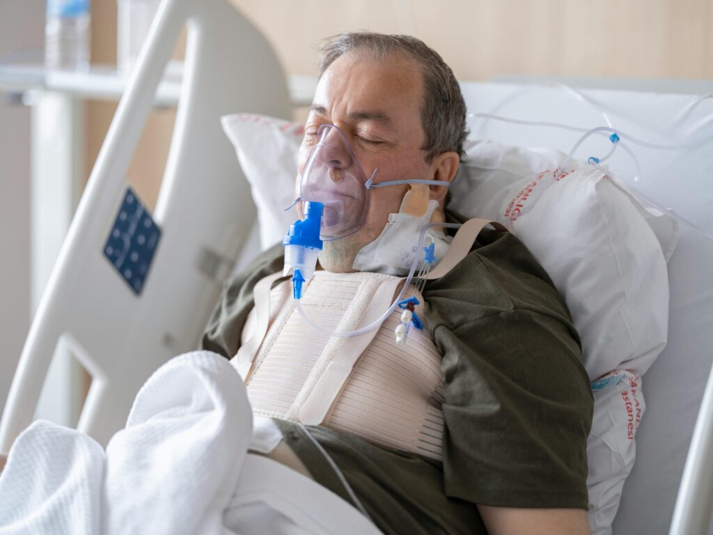 An elderly man lies in a hospital bed wearing an oxygen mask for medical care.