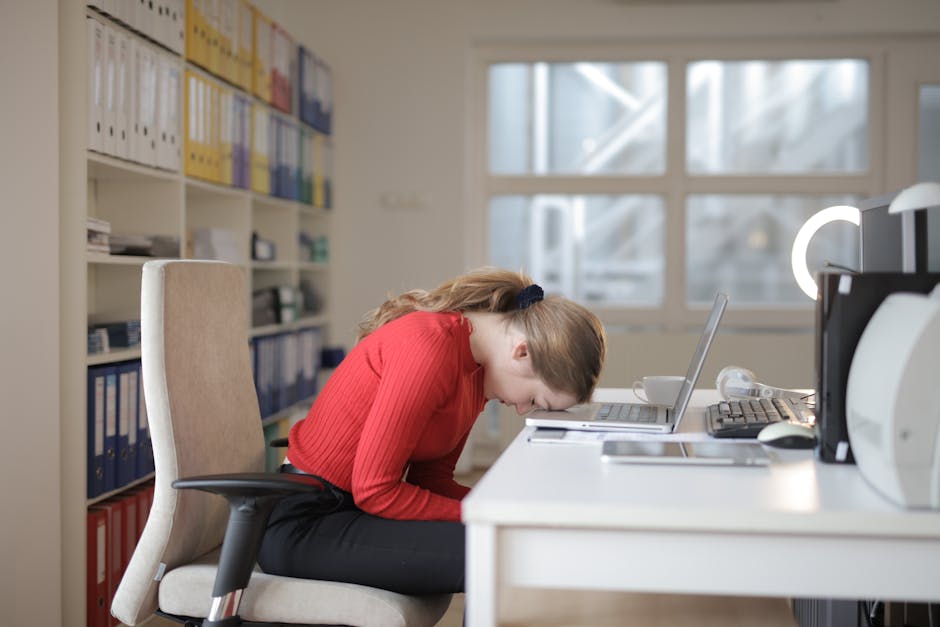 woman in burnout wearing red sweater naps on office desk beside laptop, overwhelmed by remote work.